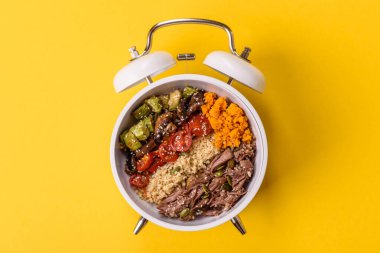 Çalar saatin arka planında kinoalı, sebzeli, etli ve yumurtalı vejetaryen salatası ya da kase. Vejetaryen yemeği yemek için kavramsal geçmiş zamanı