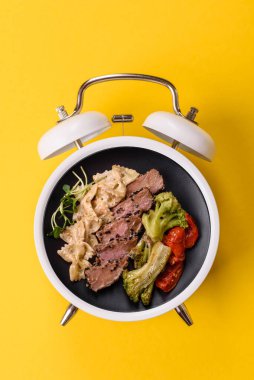 Çalar saatin arka planında et, sos ve sebzeli İtalyan makarnası. Makarna yemek için kavramsal arka plan zamanı