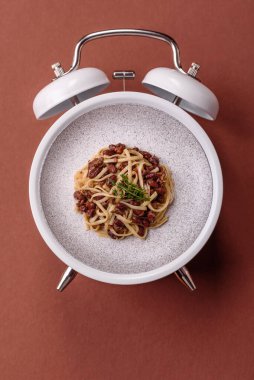 Çalar saatin arka planında et, sos ve sebzeli İtalyan makarnası. Makarna yemek için kavramsal arka plan zamanı