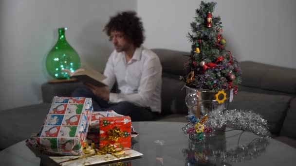 独身男性はクリスマスの日をプレゼントやクリスマスツリーと一緒に自宅で過ごす 本を読んでソファでリラックス 休日や両親のストレスや孤独はありません 自宅でのライフスタイル — ストック動画