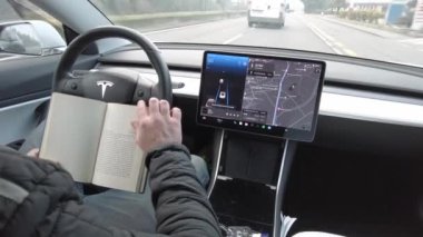 Avrupa, İtalya, Milan 12-11-2023 Tesla Electric arabasında otomatik pilot ve otomatik sürücü özellikleri - araba ilerliyor ve sürücü kitap okuyor, ellerini direksiyona koyma- destekli sürüş