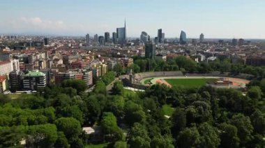 Şehir silüeti yemyeşil park ağaçlarının arkasında yükseliyor.