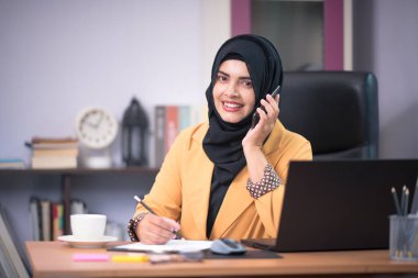 Asyalı Müslüman kadın ofis çalışanı masadaki dizüstü bilgisayarın önünde oturuyor ve ofiste mutlu bir şekilde gülümsüyor..