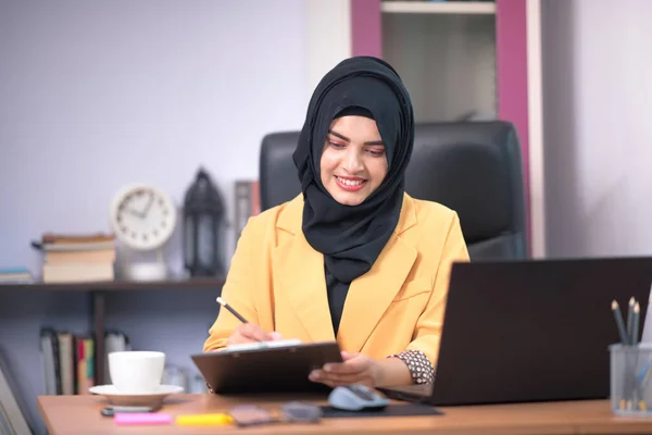 Asian Muslim woman office worker sitting in front of laptop computer at desk and smiling happy at office.