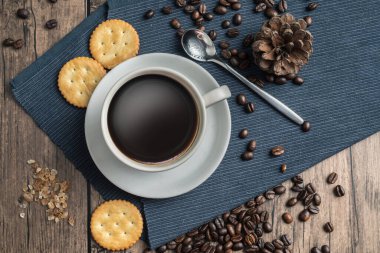 Beyaz kahve fincanındaki sıcak kahve ve etrafına bir sürü kahve çekirdeği ve sıcak, açık atmosferli, koyu arkaplanda fotokopi alanı olan ahşap bir masaya kraker..