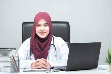 Asyalı Müslüman kadın doktor hastanın masasında oturmuş gülümseyen ve misafirperver bir yüzle kameraya bakıyordu..