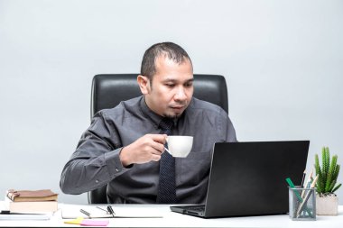 Asyalı erkek ofis çalışanı masadaki dizüstü bilgisayarın önünde oturuyor ve ofisteki bilgisayar ekranında yaptığı işi görmekten çok mutlu..