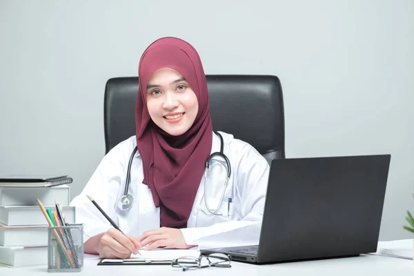 アジア系イスラム教徒の女性医師が患者のテーブルに座り 笑顔で迎えてくれるカメラを見ていた — ストック写真