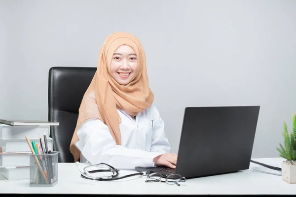 亚洲穆斯林女医生坐在病人的桌旁 面带微笑地看着摄像机 — 图库照片