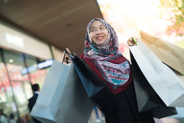 Asyalı Müslüman bir kadın alışveriş yaptığı ve mutlu yüzlü bir sürü ürün aldığı bir çanta dolusu mal taşıyor..