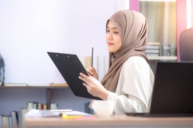 Ofis çalışanı, Asyalı Müslüman bir kadın, masada dizüstü bilgisayarın önünde oturuyor ve ofisteki dosyalara başarıyla bakıyor..