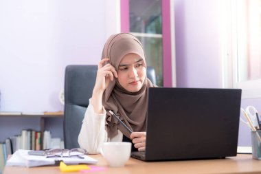 Asyalı Müslüman kadın ofis çalışanı masadaki dizüstü bilgisayarın önünde oturuyor ve ofiste mutlu bir şekilde gülümsüyor.