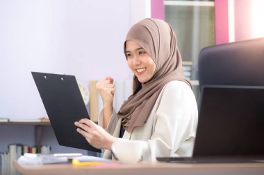 Ofis çalışanı, Asyalı Müslüman bir kadın, masada dizüstü bilgisayarın önünde oturuyor ve ofisteki dosyalara başarıyla bakıyor..