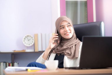 Asyalı Müslüman bir kadın için çalışan bir ofis çalışanı masasında dizüstü bilgisayarının önünde oturuyor ve cep telefonuyla konuşuyor ve ofiste çalışıyor..