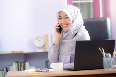 Asyalı Müslüman bir kadın için çalışan bir ofis çalışanı masasında dizüstü bilgisayarının önünde oturuyor ve cep telefonuyla konuşuyor ve ofiste çalışıyor..