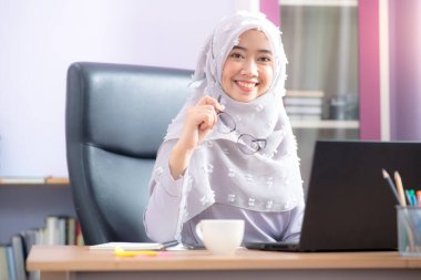 Asyalı Müslüman bir kadın için çalışan bir ofis çalışanı masasında dizüstü bilgisayarının önünde oturuyor ve cep telefonuyla konuşuyor ve ofiste çalışıyor..