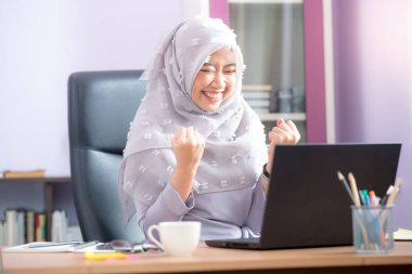 Asyalı Müslüman kadın ofis çalışanı bir masadaki dizüstü bilgisayarın önünde oturuyor ve ofisteki bilgisayar ekranında yaptığı işi görmekten çok mutlu..