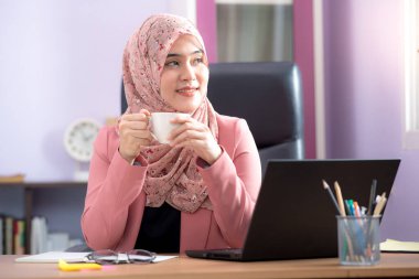Asyalı Müslüman kadın ofis çalışanı masadaki dizüstü bilgisayarın önünde oturmuş mutlu bir şekilde gülümsüyor ve elinde beyaz bir kahve fincanı tutuyordu..