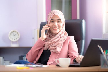 Asyalı Müslüman bir kadın için çalışan bir ofis çalışanı masasında dizüstü bilgisayarının önünde oturuyor ve cep telefonuyla konuşuyor ve ofiste çalışıyor..