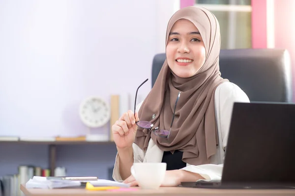 Asian Muslim woman office worker sitting in front of laptop computer at desk and smiling happy at office