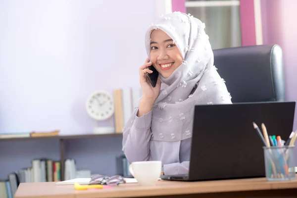 一位亚洲穆斯林妇女的办公室职员坐在办公桌前的笔记本电脑前 一边用手机聊天 一边在办公室里工作 — 图库照片