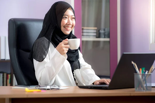 亚洲穆斯林妇女办公室职员坐在笔记本电脑前 坐在办公桌前 开心地笑着 并在办公室里拿着一个白色的咖啡杯 — 图库照片