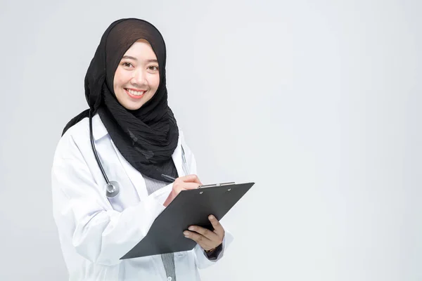 她拿着写有病历的剪贴板 拿着实验室的外套 微笑着美丽的亚裔穆斯林女医生 她正在白色背景下看着相机 免版税图库图片