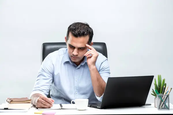 An Asian office worker is sitting in front of a laptop computer at his desk and is having a headache and is not happy to see the work with his documents at the office.