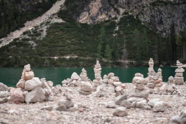 A tranquil natural scene showcasing a collection of balanced stone cairns on a rocky shore, with a pristine turquoise mountain lake and lush greenery in the background. The serene environment embodies harmony and peacefulness. clipart