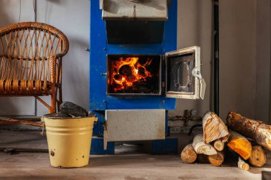 Açık kapısı olan mavi, katı bir yakıt kazanı içinde yanan kütükler olduğunu ve sıcak bir parıltı yaydığını gösteriyor. Kesilmiş odun yığını yere dizildi. Sarı metal bir kovada kömür parçaları var..