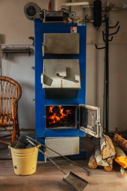 Açık kapısı olan mavi, katı bir yakıt kazanı içinde yanan kütükler olduğunu ve sıcak bir parıltı yaydığını gösteriyor. Kesilmiş odun yığını yere dizildi. Sarı metal bir kovada kömür parçaları var..