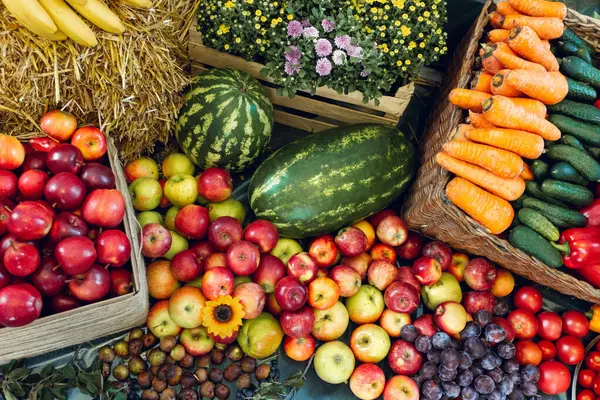 Taze sebzeler, meyveler, çiçekler, karpuzlar saman üzerinde, sandıklar ve sepetler içinde ve karanlık bir yüzey üzerinde dizilmiş.