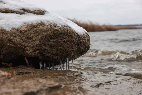 Kıyıda buz saçakları olan, dalgaların çarptığı, yüzeyinin bazı kısımlarında kar bulunan, kış soğuk atmosferi olan bir kayanın yakın çekimi.