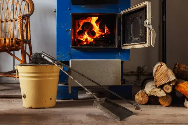 Açık kapısı olan mavi, katı bir yakıt kazanı içinde yanan kütükler olduğunu ve sıcak bir parıltı yaydığını gösteriyor. Kesilmiş odun yığını yere dizildi. Sarı metal bir kovada kömür parçaları var..