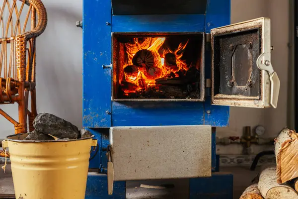 Açık kapısı olan mavi, katı bir yakıt kazanı içinde yanan kütükler olduğunu ve sıcak bir parıltı yaydığını gösteriyor. Kesilmiş odun yığını yere dizildi. Sarı metal bir kovada kömür parçaları var..