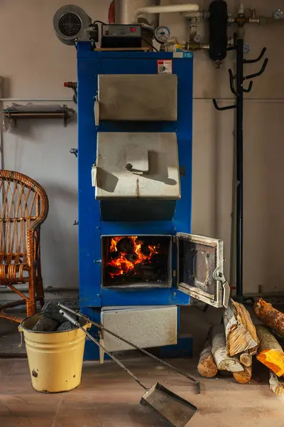 Açık kapısı olan mavi, katı bir yakıt kazanı içinde yanan kütükler olduğunu ve sıcak bir parıltı yaydığını gösteriyor. Kesilmiş odun yığını yere dizildi. Sarı metal bir kovada kömür parçaları var..