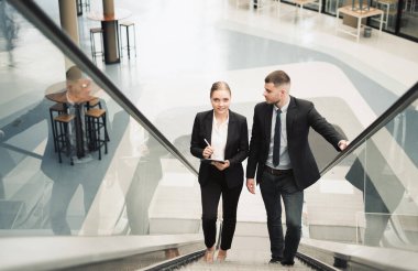 Genç işadamı ofiste yürüyen merdivene tırmanırken kameraya bakıyor.