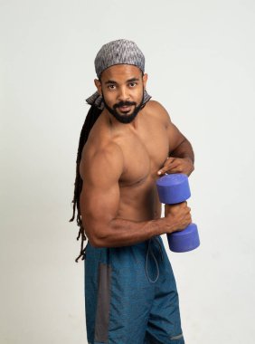Rastalı ve sakallı atlet kaslı latin adam. Kolunu bir dambıl tutarak kameraya bakıyor. Beyaz arka planda izole halterlerle egzersiz yapıyor.
