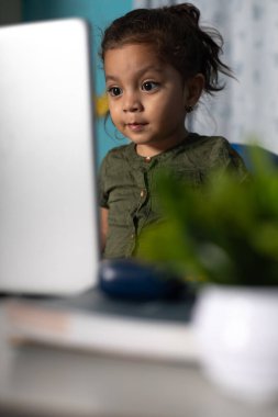 Beautiful 2-year-old Mexican girl, in the background of the photo, looking very attentively at the laptop clipart