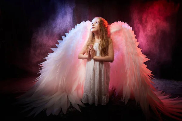 A girl in a white shirt, wings and with long hair, looking like a angel. A young model posing at a photo shoot with smoke, fog, flour, dust. A little fairy girl on dark black background