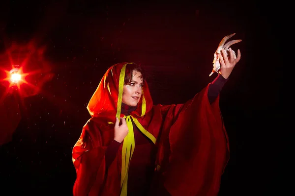 Witch woman in red cloak with skull on black background and light in night. Female warrior in Dark