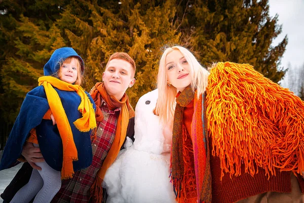 Happy parents and child gathering in snow-covered park together sculpting funny snowman from snow. Father, mother and kid daughter playing outdoor in winter forest. Family active holiday