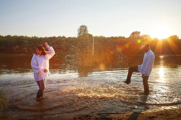 Water spray, partial focus. Beautiful adult couple has fun in nature in water in river or lake in the summer evening at sunset. Guy and girl swim and relax outdoor in clothes in white shirts and jeans