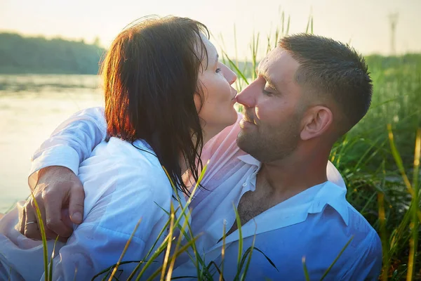 Beautiful adult couple having hugs and fun on nature in the water of a river or lake in the summer evening at sunset. A guy and a girl swim and relax outdoors in clothes in white shirts and jeans