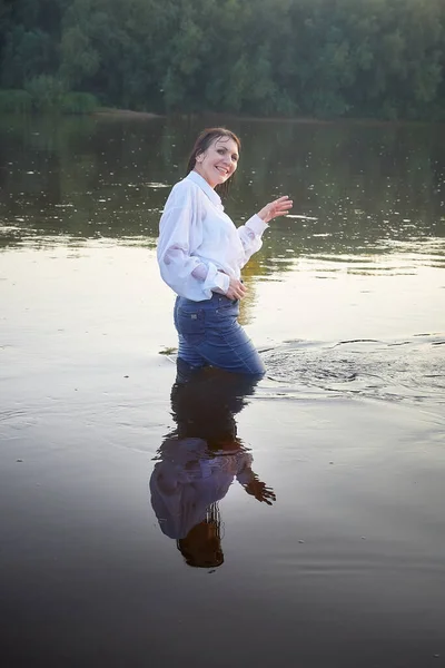 Portrait of beautiful young woman in stylish dress with white shirt and jeans in water of river or lake in nature landscape in summer evening