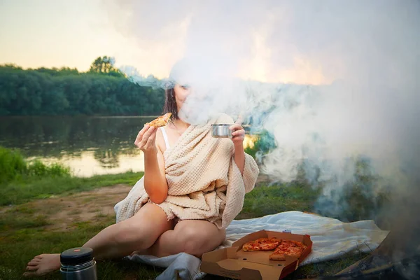 A wet girl in a light blanket after bathing with a piece of pizza on the bank of a river or lake on summer evening near a campfire with smoke and fog. Nice summer evening