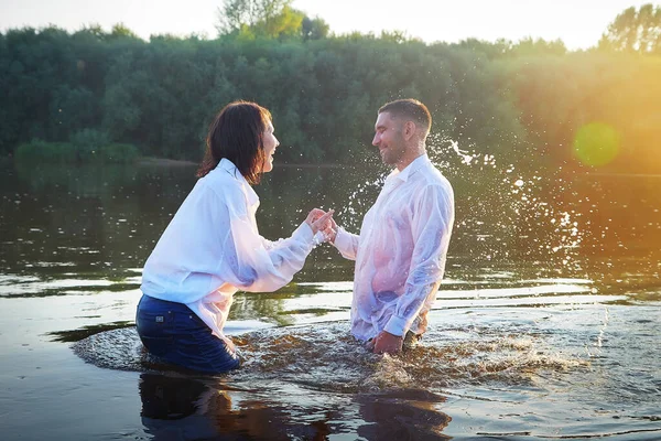 Beautiful adult couple has fun in nature in the water in a river or lake in the summer evening at sunset. A guy and a girl swim and relax outdoors in clothes in white shirts and jeans