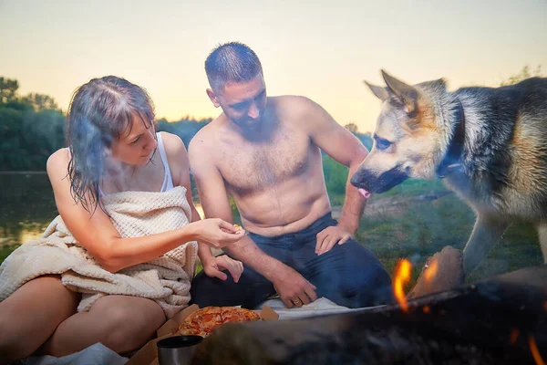 Happy wet couple relaxin, having fun and hugs with big dog near fire in camping on nature in summer sunny evening in sunset. Family or lovers have date and rest outdoor. Concept of love