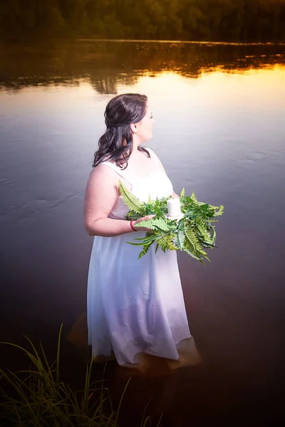 Slavic plump plump chubby girl in long white dress on the feast of Ivan Kupala with flowers and water in a river or lake on summer evening