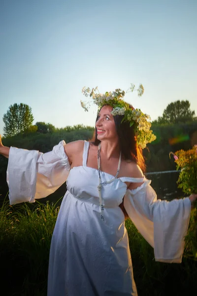 Portrait of adult mature brunette woman in a white dress, sundress and wreath of flowers in summer in the evening at sunset. Celebration of the Slavic pagan holiday of Ivan Kupala
